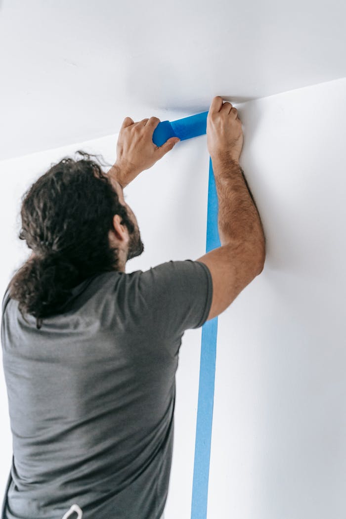 About A man applying blue tape on a wall during a home renovation project, showcasing painting preparation.